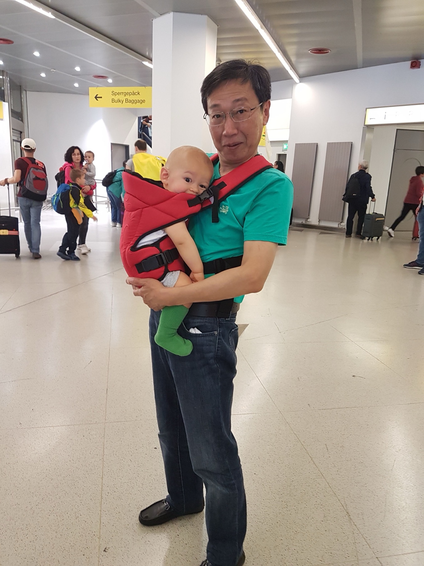 Mio suocero porta mio figlio di un anno in un marsupio rosso in aeroporto dopo un volo in famiglia.