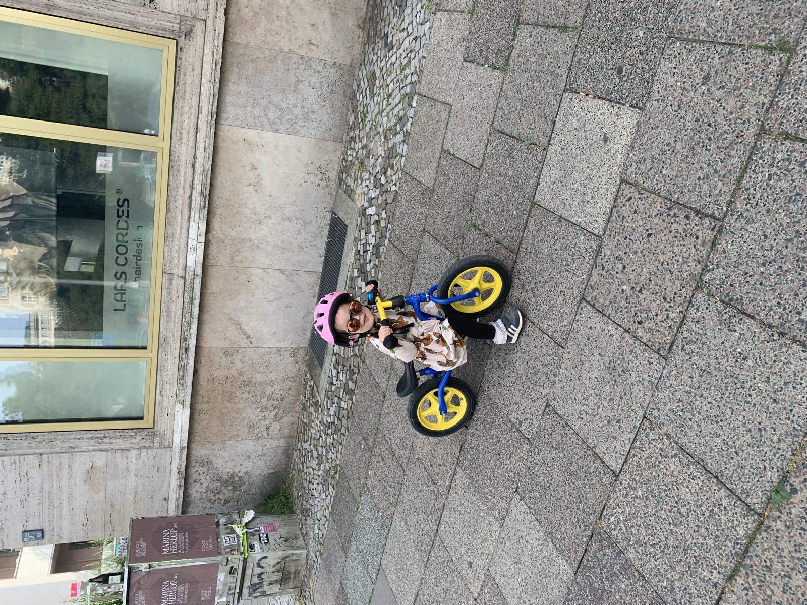 Meine Tochter sitzt auf demselben blau-gelben Laufrad auf einem Stadtgehweg.