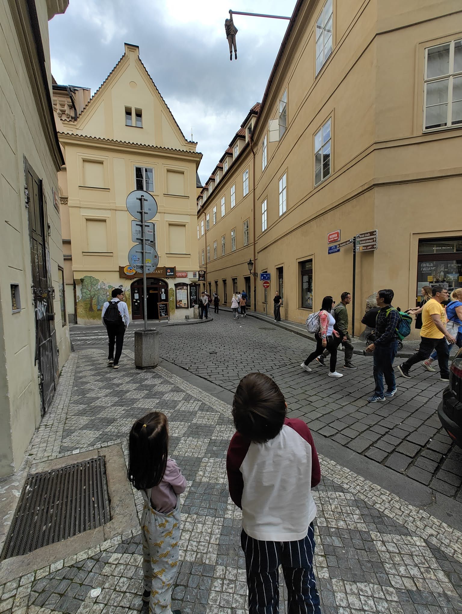 The children walking through a quiet Prague lane below the hanging figure sculpture, a small surreal stop between old streets.